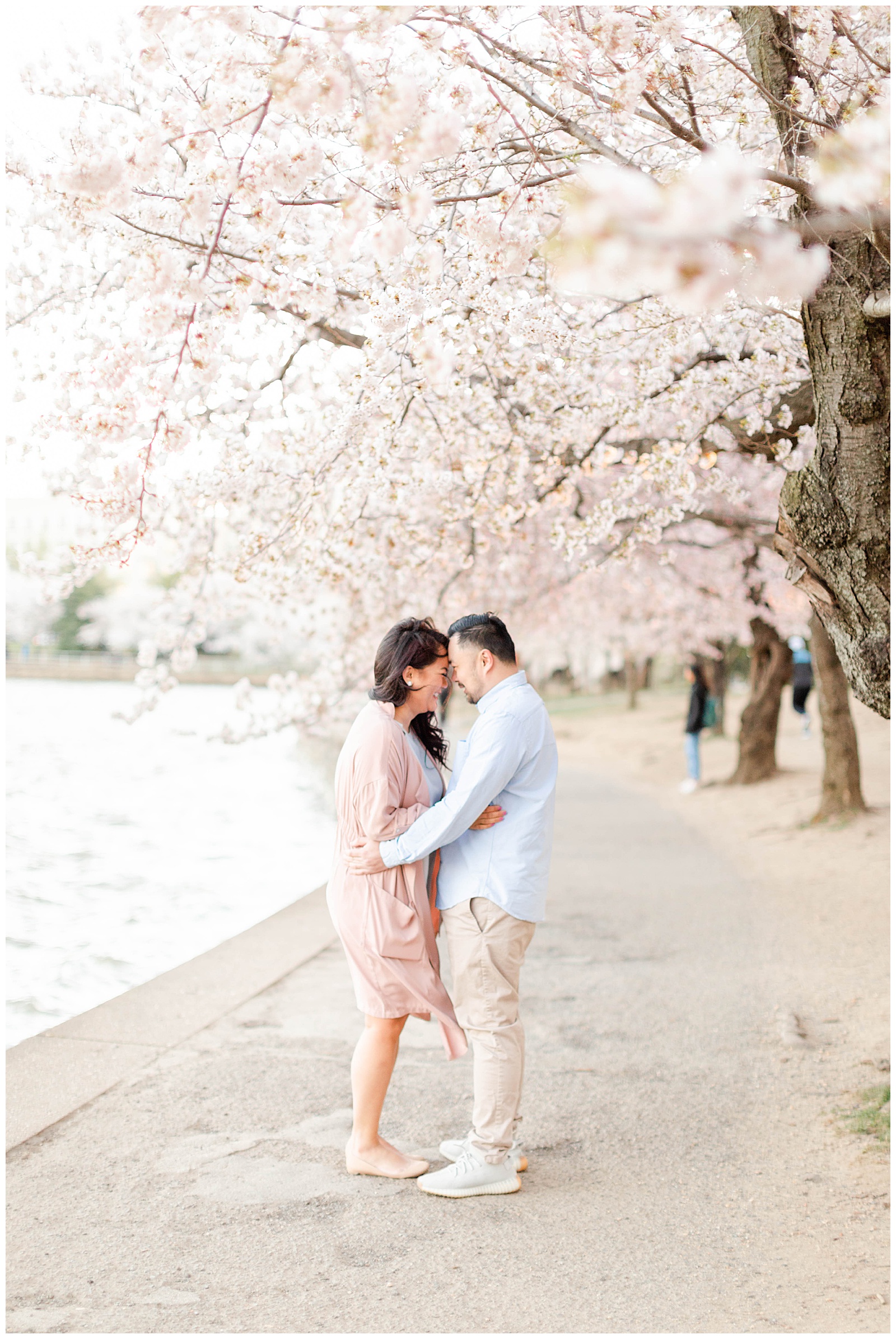 Couple standing next to Tidal Basin in Washington D.C with cherry blossom trees blooming in background.